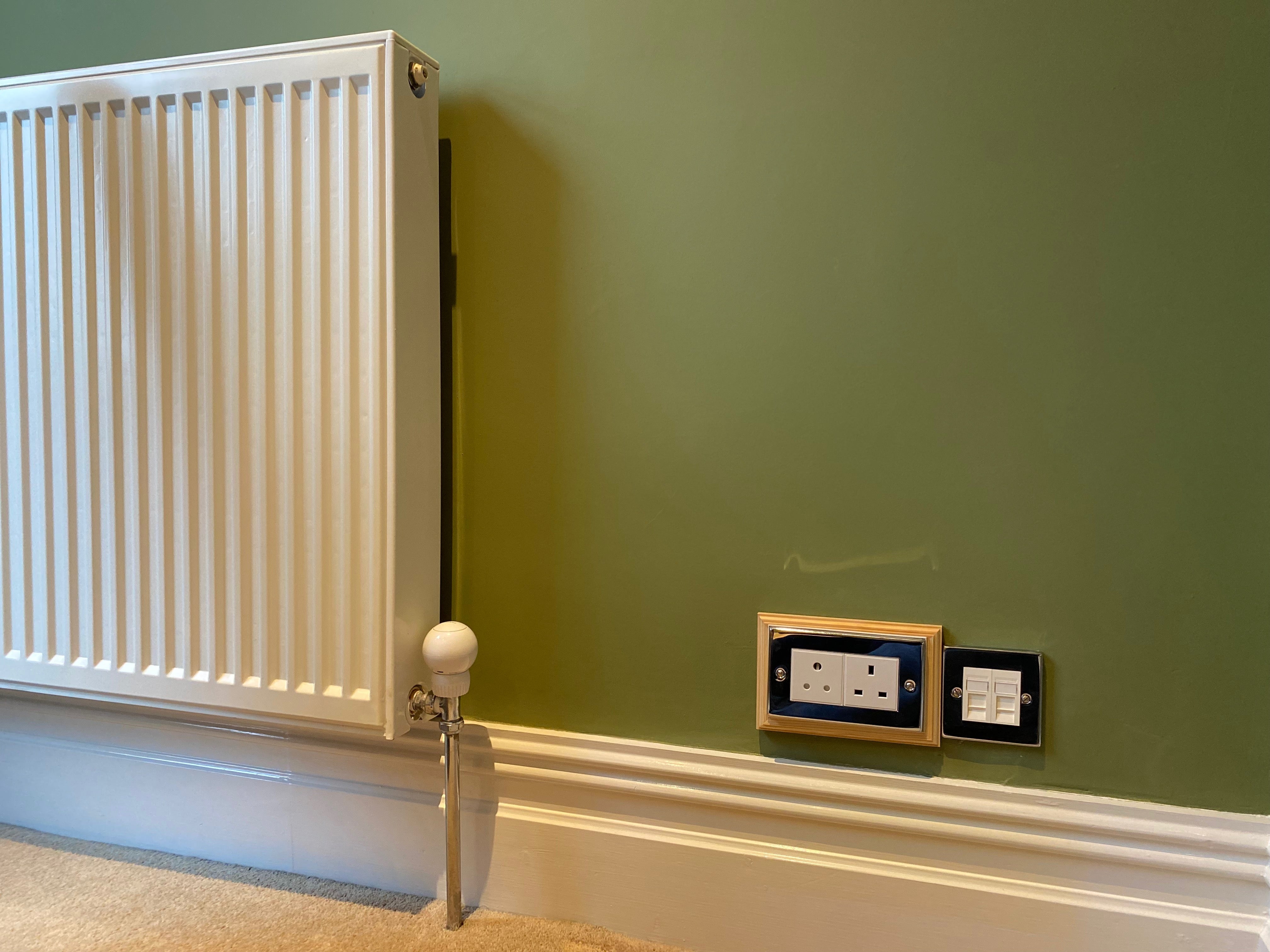 A radiator with a smart FIBARO Heat Controller on a carpeted floor against a green wall, beside a plug socket.