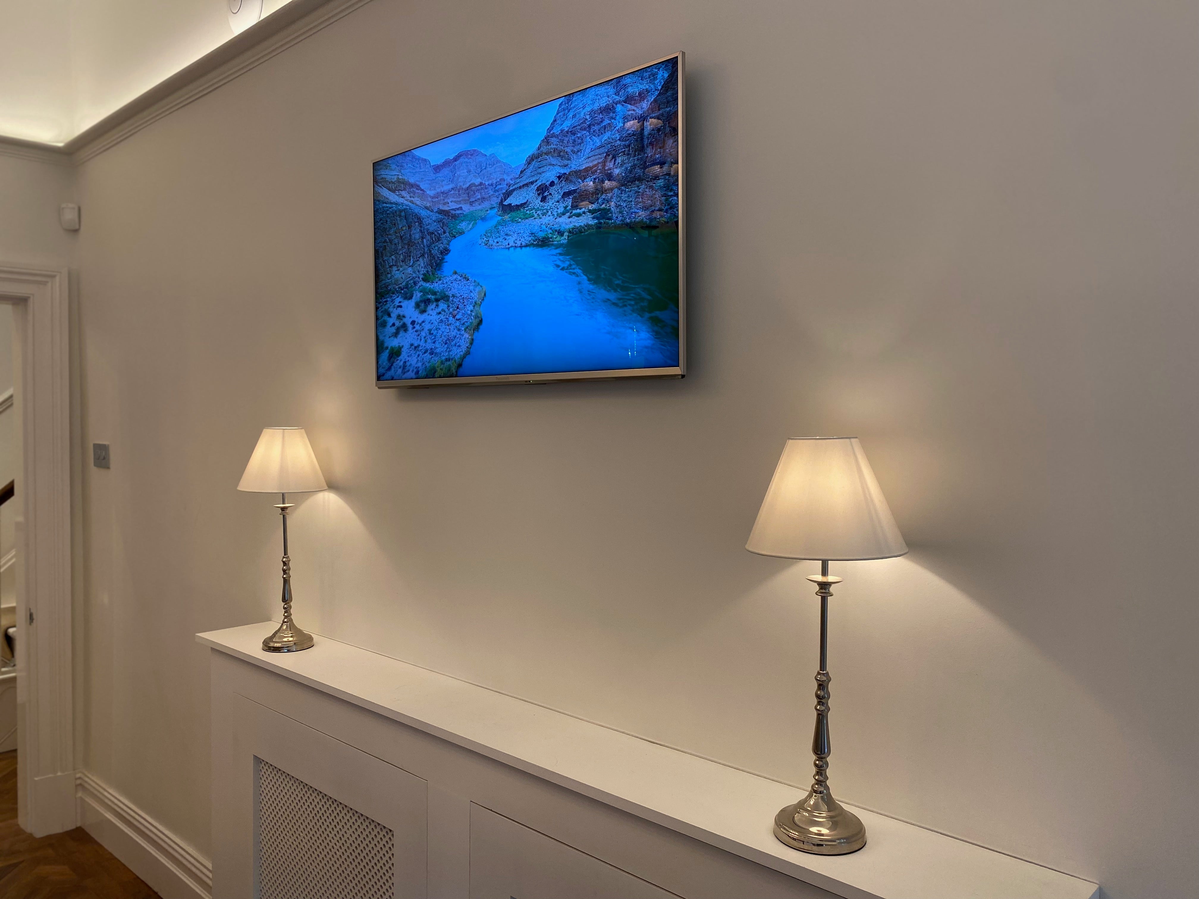 A wall mounted TV displays an image of a river, with two metal lamps underneath. 