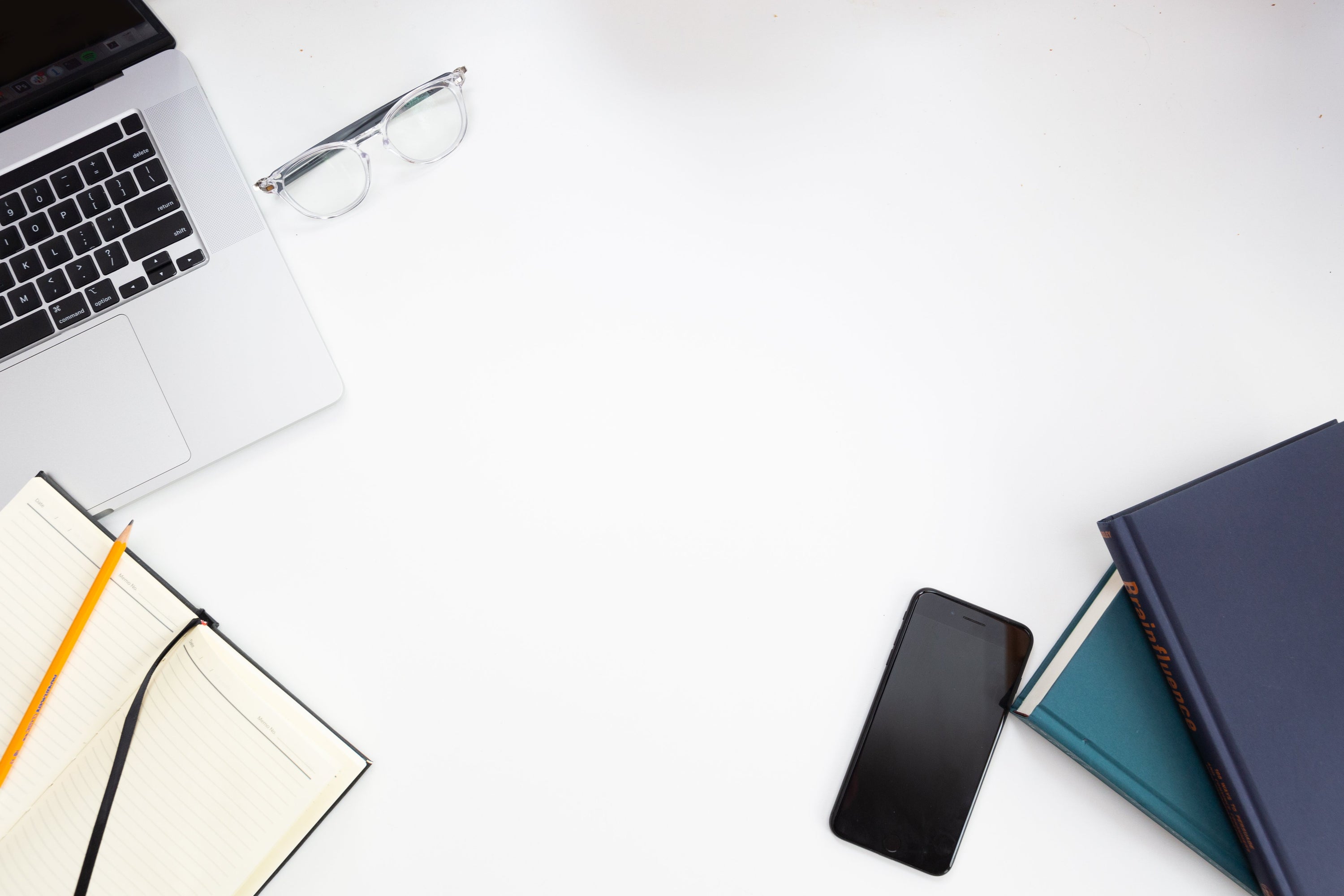 A laptop, a smartphone, a pair of reading glasses and several books sit on top of a white desk, seen from above. 